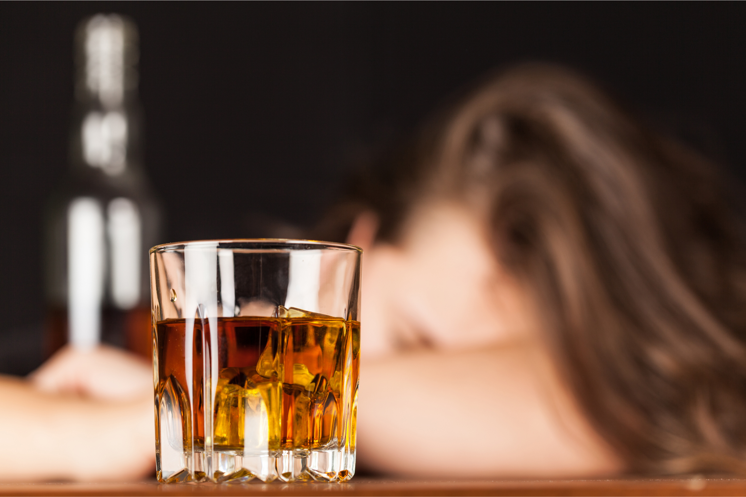 woman sitting with her head on the table next to a bottom and glass of whiskey showing signs that someone is an alcoholic